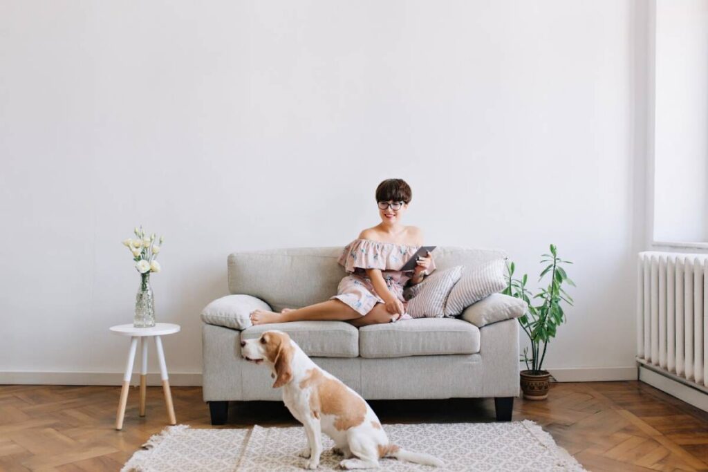 Woman in off-shoulder dress sitting on a sofa using a tablet, with a dog beside her and a vase of flowers nearby.