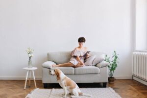 Woman in off-shoulder dress sitting on a sofa using a tablet, with a dog beside her and a vase of flowers nearby.