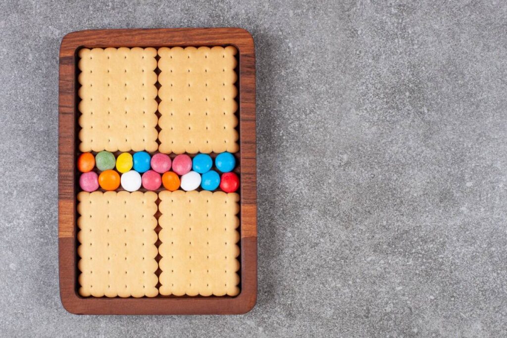 Rectangular wooden tray filled with neatly arranged biscuits and a row of colourful candies on a gray surface.