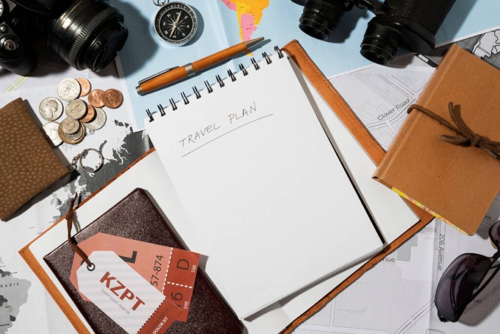 Open notebook with "TRAVEL PLAN" written, surrounded by a camera, coins, a compass, maps, and a passport on a table.
