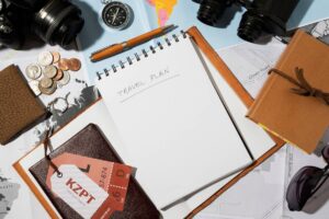 Open notebook with "TRAVEL PLAN" written, surrounded by a camera, coins, a compass, maps, and a passport on a table.