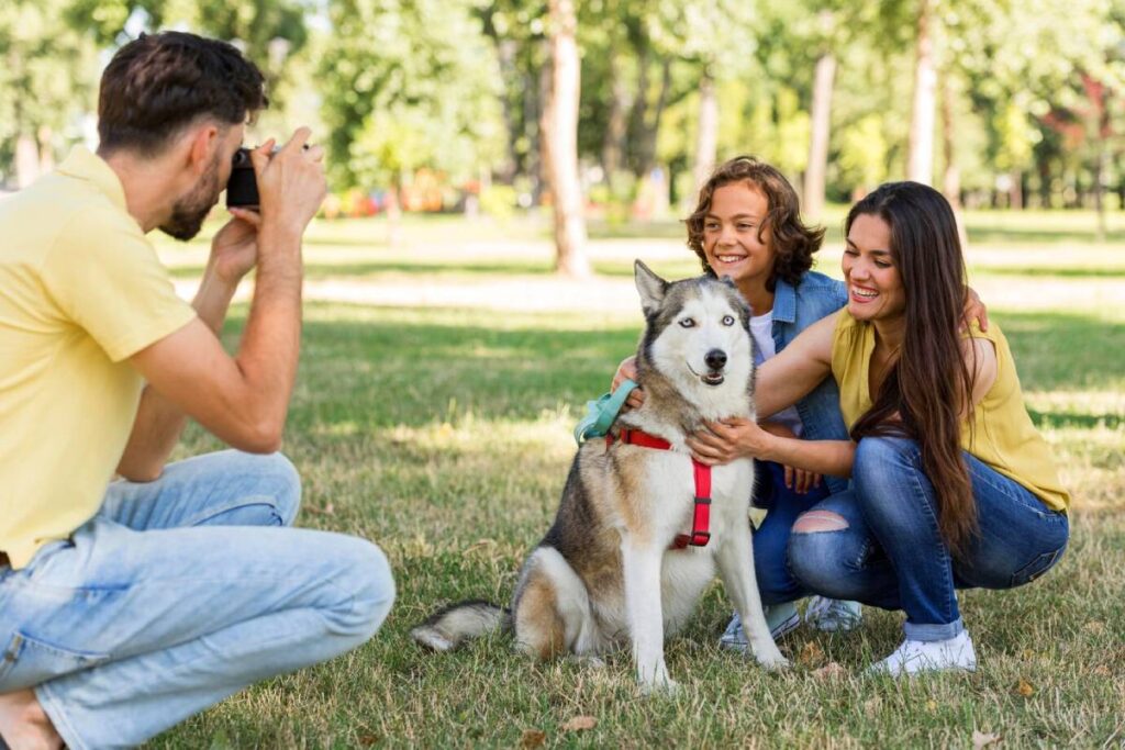 Family enjoying time in park with husky dog, while man takes their photo.