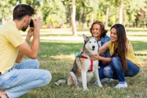 Family enjoying time in park with husky dog, while man takes their photo.