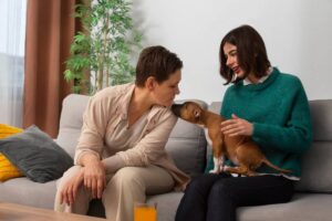 Two women on a couch; one kisses a small dog held by the other in a cozy living room.
