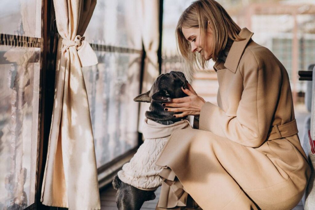 A woman in a beige coat affectionately cuddling a black French Bulldog wearing a sweater, sitting near a window.