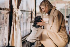 A woman in a beige coat affectionately cuddling a black French Bulldog wearing a sweater, sitting near a window.