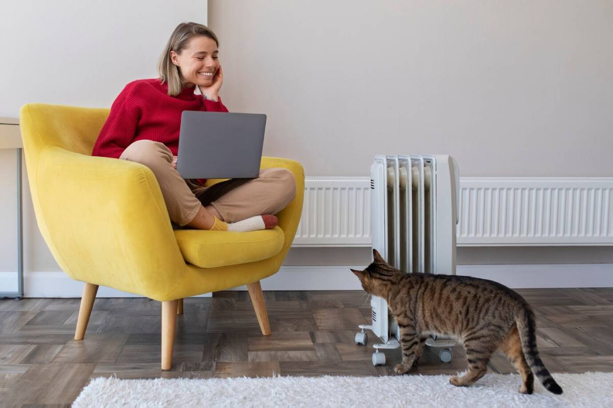  A woman smiling, working on a laptop in a yellow armchair, with a cat beside her near a radiator.