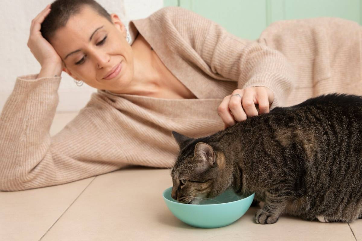  Woman in a beige sweater smiling at a tabby cat eating from a blue bowl.